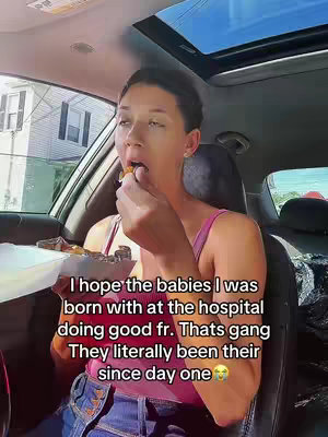 A woman is sitting in the driver's seat of a car, holding a container of food up to her mouth. She's wearing a pink top and has short dark hair. The background shows a residential street scene and the interior of the car.