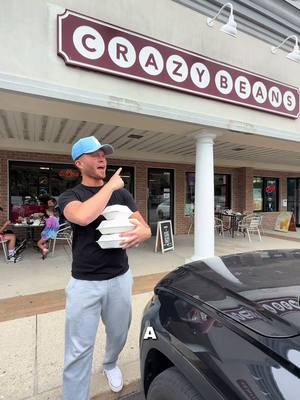 A man in a blue cap, black t-shirt, and gray sweatpants stands outside a building with a 'CRAZY BEANS' sign, holding white containers and pointing with a raised finger. A black vehicle is parked nearby. Other individuals are seen seated at tables in the background.
