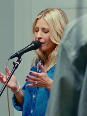 The image captures a woman singing into a microphone, with her eyes closed in apparent emotion or concentration. She's wearing a blue denim shirt and her blonde hair is styled. Beside her, the shoulder and arm of another person is seen.
