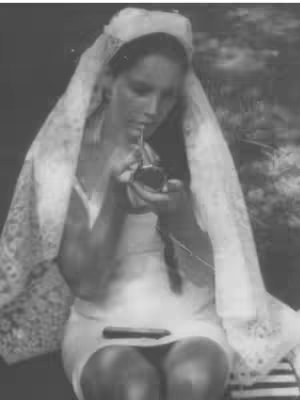 A black and white image features a woman, likely in her youth, dressed in what appears to be a white sleeveless dress or slip. She is wearing a veil and holding a small, dark object, possibly a prop like a tobacco holder or a decorative item, near her mouth, as if she is about to bring it to her mouth or is already using it. The image is softly lit with many grey tones. There is a blurry background.