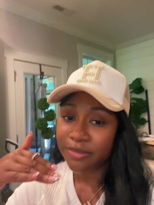 A selfie-style shot features a woman in a room. she faces the camera with a seemingly neutral expression. Her complexion appears natural, and her hair is black and parted on the side. She wears a white and tan trucker hat with the initial "H" on the front, a necklace, and a ring. The background includes a view into another room and appears to be indoors under natural lighting.