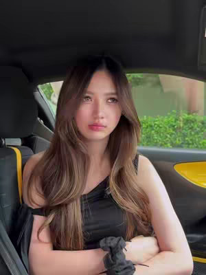 A young woman with long brown hair is seated inside a car. She's wearing a black top and has her arms crossed. The car's interior is dimly lit, with the focus on the woman's face and upper body, indicating a shot taken inside the car.