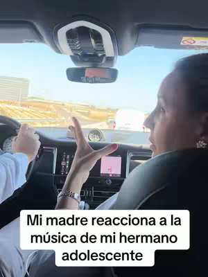 Interior view from inside a vehicle. A woman in the front seat turns towards the side, her expression animated with raised hands. A hand is visible on the steering wheel. The background shows a road and buildings.