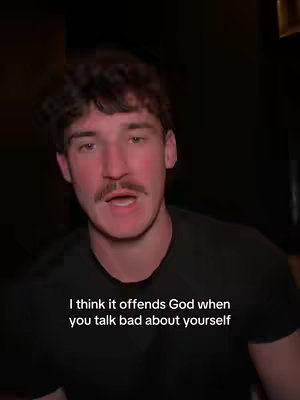 A man with dark hair and a mustache is centered in the frame, speaking directly to the camera. The background is dimly lit. Text is overlaid on the bottom of the frame.