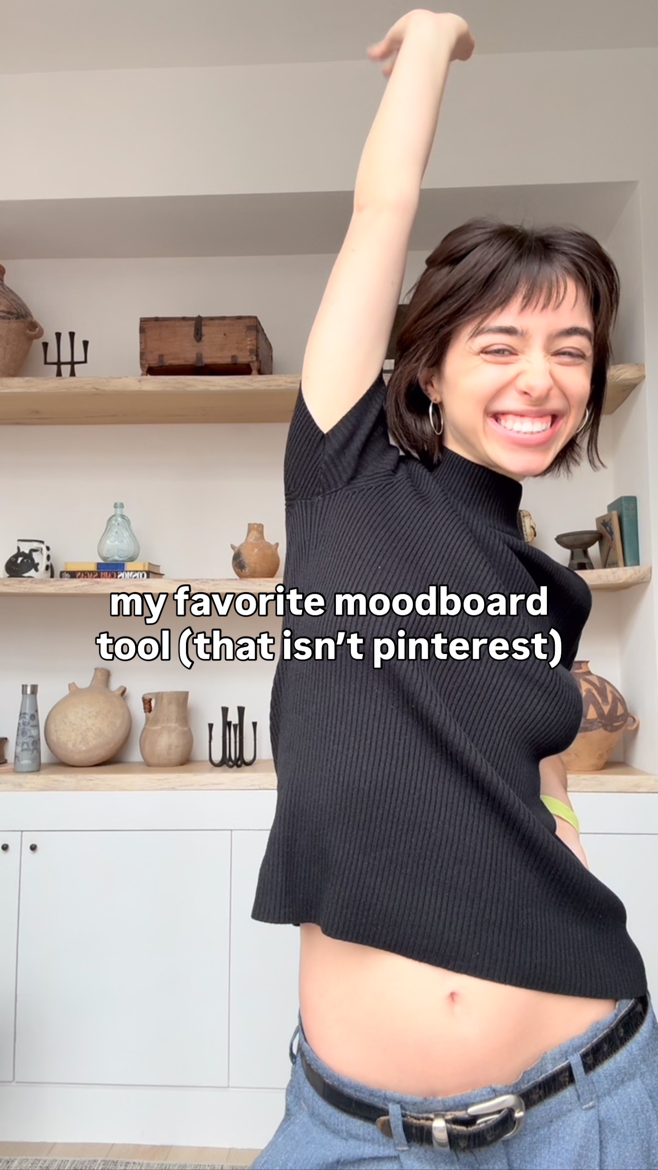 A woman is smiling widely, holding her right arm up in the air. She is wearing a black, ribbed top, and light wash denim pants with a black belt. The background includes white cabinets and wooden shelves with various items.