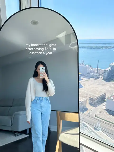 A woman is standing in front of a large mirror, holding a phone up to take a selfie. The mirror reflects her and the room, which includes a couch to the left and a view of the city outside a window to the right. Text is overlaid on the mirror.