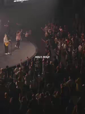 A vertical shot of a worship service in a large hall. The focus is on a stage where musicians and singers are performing. The crowd fills much of the space, with many people raising their hands in worship. The lighting is dim, with spotlights on the stage creating a sense of spiritual atmosphere. The text overlay 'HOLY, HOLY' is centered at the bottom of the screen.