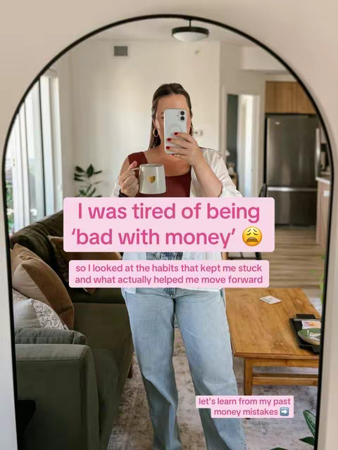Selfie taken in front of a large arched mirror reflecting a room. The frame houses a woman holding a white mug with a gold heart design. She is wearing a white blazer, a light-colored top, and light-wash jeans. Text overlays in pink boxes add information. The background reveals a couch and a wooden table creating a warm, inviting atmosphere.