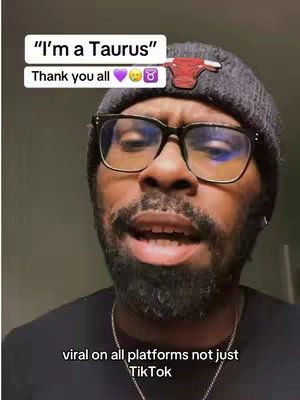 A close-up shot features a Black man with a short beard and glasses, speaking towards the camera. Above him, two white rectangular boxes display text overlays. The upper box reads 'I'm a Taurus', while the lower box says 'Thank you all' followed by emojis including a purple heart and the Taurus zodiac symbol. He is wearing a black beanie with a bull emblem, and the background is a plain, off-white wall.