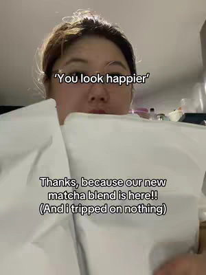 A person is holding up a large white container, with text layered in front of them. The background is a simple, indoor setting. The person's face is visible, and they appear to be smiling. The text overlay has two lines, the first saying "You look happier", the second saying "Thanks, because our new matcha blend is here!!".