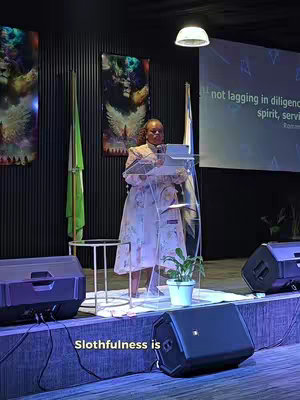 The cover image shows a woman standing behind a transparent podium, speaking into a microphone. She is on a stage with sound equipment, a small plant, and a banner. A screen in the background displays a quote. The lighting is dim with blue and green hues. Overall the image is composed of a professional setup for speaking or giving a sermon.