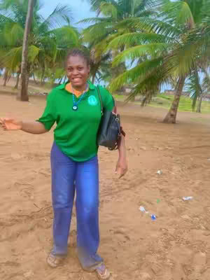 A woman stands on a sandy beach, near palm trees. She is wearing a green shirt and blue jeans, and the sky is overcast. The background includes palm trees, and the person is positioned in the front.
