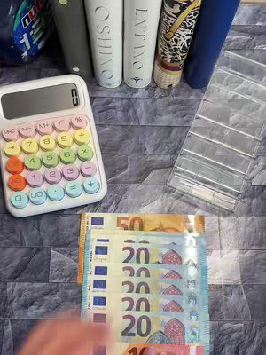 A top-down shot shows a collection of items on a textured, stone-like surface. A white calculator with colorful buttons, a stack of euro bills, and a stack of envelopes. Several cosmetic tubes and bottles are arranged in the background. A hand is partially visible at the bottom, near the money.