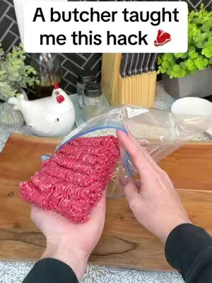 The image shows hands holding a package of raw ground meat ready to be placed into a plastic zip bag. The ground meat is red and seems to be stored in the freezer. In the background, there are kitchen utensils and decorations.