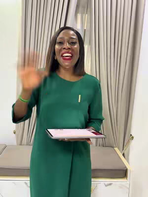 A woman stands in a well-lit room, smiling and waving at the camera. She's wearing a green dress and holding a tablet. Soft-toned curtains are in the background.