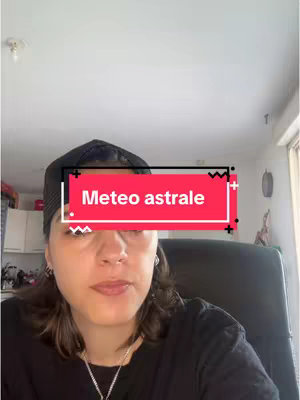 A woman with dark hair is centered in the frame. She is wearing a black shirt and a dark cap worn backwards. A text overlay, a rectangle with the words "Meteo astrale" in white letters, is on top of her head in a red background. The room is blurred behind her, with some cabinets on the left and a window on the right.