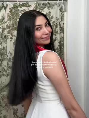 A woman with long, dark hair gazes at the camera with a slight smile. She wears a white top with a red trim. The background shows a white curtain with a floral design. The text overlayed states, "la suerte se pierde el sueno por eso estando vuelta a la suenos es suerte".