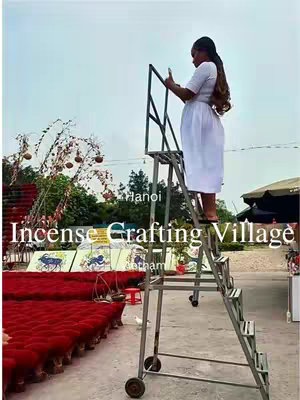 A woman in a white dress is standing on a metal ladder holding a phone, taking a photo. The background features a cloudy sky, greenery, and stacks of red incense.