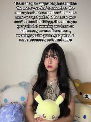 A well-lit selfie featuring a young woman with long, dark hair and bangs, fair skin, and pinkish makeup. She is holding a Pikachu plush and is surrounded by other stuffed animals in a soft background. The overall composition is warm and friendly, with a focus on the woman and the plush.