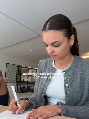 A woman is sitting at a desk, writing with a pen on a piece of paper. She is wearing a gray cardigan over a white top, and her dark hair is pulled back in a high ponytail. Text overlayed on the image reads: 'sometimes it's 0% motivation but 100% discipline.' The background includes a shelf with books and decorative items. The overall composition gives a vibe of studying.