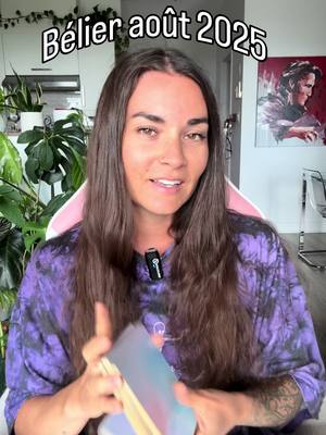 A woman with long brown hair wearing a purple tie-dye shirt is looking at the camera and smiling. She is holding a deck of tarot cards in her hands. The background includes a glimpse of a white kitchen and a painting.