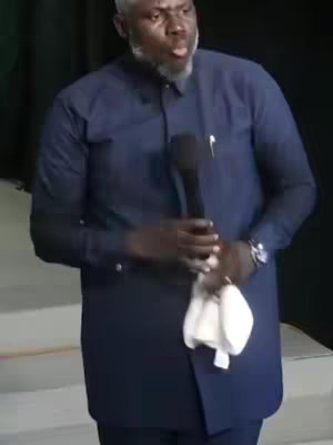 A man in a navy blue suit and a white cloth in his hands speaks into a microphone. The background is a neutral color with a slight off-white wall, and the lighting appears to be natural or soft.