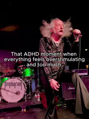 A man with white, frizzy hair is on a stage with a microphone, looking up and to the side with a wide-eyed expression. The scene suggests a performance or concert, with a drum set slightly behind him. Text overlayed at the bottom mentions ADHD and overstimulation.