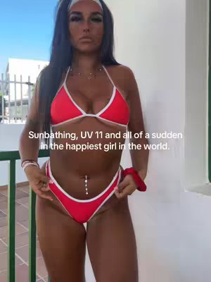 A woman in a red bikini stands on a balcony, holding the straps of the bikini bottom. The background includes a bright blue sky and green plants. Text overlayed in white on the image describes her mood after sunbathing.