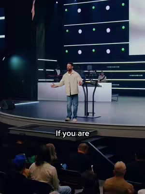 A man dressed in a light-colored jacket and jeans is speaking onstage with his arms outstretched. The stage is lit with cool lighting and has a screen background with green accents. Foreground text reads 'If you are'. Audience seating is visible at the bottom.