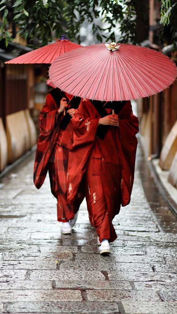 Two figures walk along a wet, stone path in an alleyway. Both are wearing long red robes, with white shoes visible. They each hold a large, red umbrella with a detailed design. The background consists of buildings with a slight blur.