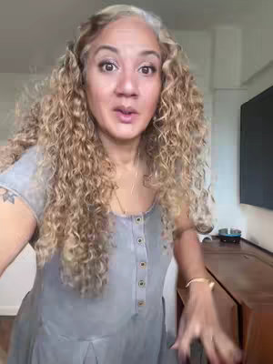 A woman with curly blonde hair is shown from the chest up in a selfie-style shot. She is looking directly at the camera with a slight smile. The background is a nondescript room.
