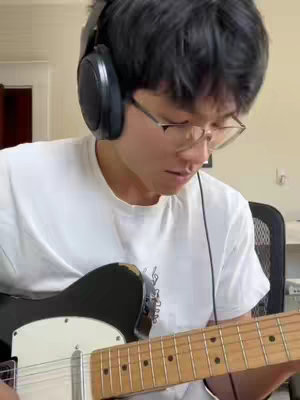 A person with glasses is playing a black electric guitar while wearing headphones, the background is a light-colored room.