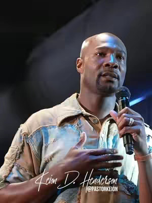 A medium shot of a Black man speaking into a microphone and wearing a denim shirt, with dim, blue-toned stage lighting in the background. Text in the lower left corner reads 'Keion D. Henderson' with '@pastorKeion' below.