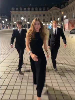 A woman in a black dress walks towards the camera, flanked by two men in suits. They are on a paved area at night, with buildings and streetlights in the background.