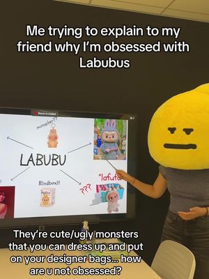 A person wearing a yellow lemon-shaped head covering points to a presentation screen showing Labubu toy illustrations, diagram with text, and a white table in front of a plain black wall.