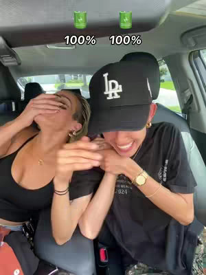 Two women are seated in a car, possibly in the front seats. One woman is covering her face with her hand while the other woman smiles with the left arm wrapped around around an arm on the face-covered woman. Above their heads, there are two green energy drink cans and texts saying '100%' for each.