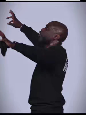 A man with a shaved head and a short beard is shown from the chest up, with his arms raised and face upturned. The background is a plain, solid light color. He is wearing a black long-sleeved shirt with some text on the back. The man's skin has a sheen of sweat.
