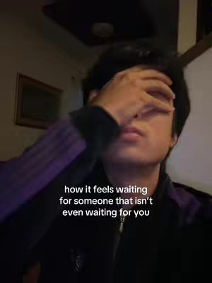 The image is a selfie of a person, seen from the chest up, with their right hand covering their eyes, suggesting sadness or frustration. The background is dimly lit, with some visible objects to the left. Text overlayed on the image expresses a sentiment about waiting for someone. The person wears a jacket that is black and purple.