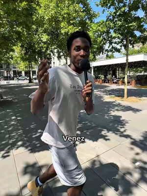A young man runs towards the camera holding a microphone, gesturing with his other hand. He wears a light-colored t-shirt, shorts, and sneakers. The background shows a sunny outdoor scene with trees and a building.