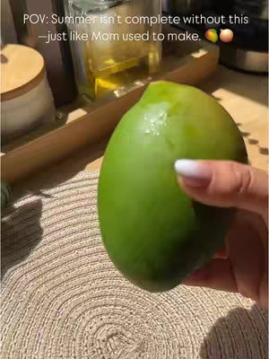 A hand holding a large green mango is the focal point, set against a kitchen or countertop background. The hand, with manicured nails, grasps the mango near its center. In the upper left corner, a wooden cutting board and a partially visible jar, likely holding oil, rest. The background appears to be a light-colored countertop with a woven placemat. Text overlays the image, suggesting the scene is a POV shot. The overall composition has a natural, inviting, and homey feel.