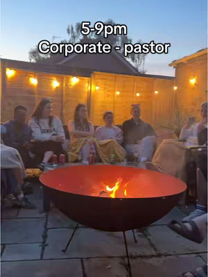 The cover image shows a backyard scene at dusk. A group of people are gathered around a fire pit. The fire pit, a large metal bowl, is the focal point, with flames visible. String lights are strung across a wooden fence in the background, and a house is partially visible. The people are sitting on benches or chairs, some wrapped in blankets. The overall composition is warm and inviting.