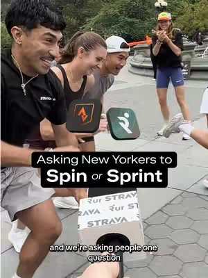 A group of four people in an urban setting, presumably during an event. A man on the left is smiling, wearing a necklace and a black top with white shorts. Beside him, a woman and another man are laughing, while in the background, a woman in a hat holds an object (likely the mic, which is not visible) and a box with a runna/strava logo on the front. Overlay text asks "Asking New Yorkers to Spin or Sprint". The overall vibe is energetic and interactive.