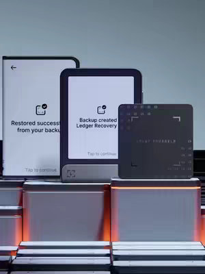 The cover image features three digital devices: a rectangular dark screen with the text "TRUST YOURSELF" on the right, flanked by two other devices with white screens displaying texts like "Restored success from your backup" and "Backup created Ledger Recovery." All devices are positioned on a three-step metallic platform.