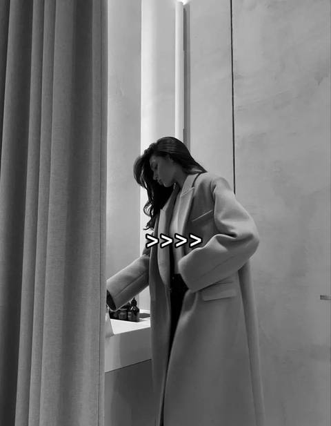 A grayscale image shows a woman in a long coat, seen from the side, with long dark hair. She leans slightly, her head down as if looking at something. A text overlay of chevrons (>>>) appears near the bottom of her coat, possibly as a video hook. The background looks like a minimalistic interior setting, featuring drapes to the left and a wall or partition on the right.