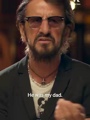 A medium shot of a man with gray hair and a beard wearing dark, thick-rimmed glasses. He's dressed in a dark blazer over a white t-shirt with a graphic on it, wearing a silver necklace with a pendant. Below the man, in white text against a background is the text: "He was my dad."
