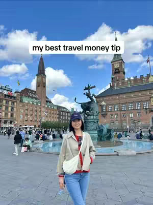 A woman stands in front of a large building with a clock tower, a fountain, and many people in the background. Above her head is a text overlay which reads "my best travel money tip". She is smiling and wearing a hat.