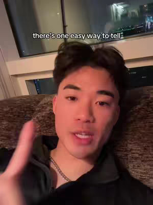 A young man with short black hair is seated on a brown couch with a dark background, the man is facing the camera and appears to be talking. A text overlay reads 'there's one easy way to tell'.