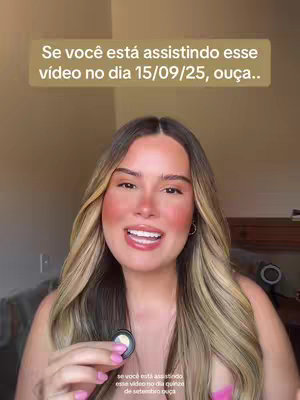 A woman with blonde hair and rosy cheeks smiles. She faces the camera, holding a small object. The background is a blurred interior with soft lighting.