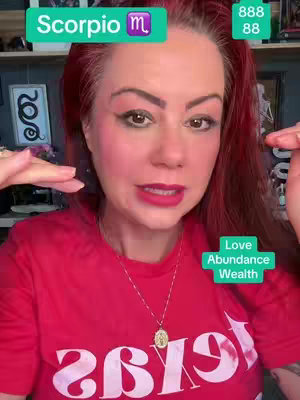 A woman with red hair and bright red lipstick faces the camera. Text overlays include her astrological sign, a number, and a callout of "Love, Abundance, Wealth". The woman is wearing a red t-shirt that has a white design on it, and a silver cross necklace.