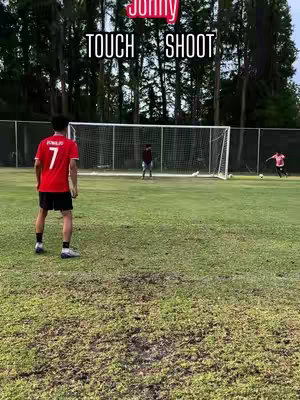 A soccer field on a sunny day. In the foreground, a person in a red soccer jersey with the number 7 on the back stands facing away from camera. Text overlay suggests a soccer challenge. In the background, two additional players are present with the net.